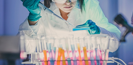 A laboratory worker holding a pipette and a Petri dish, behind a row of test tubes